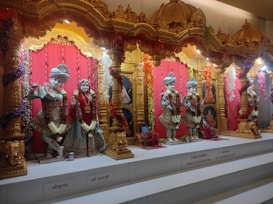 Several elaborately dressed statues are displayed in a temple setting with gold embroidered clothing and adorned with colorful garlands. The background features ornate golden arches and rich red curtains. The setting is richly decorated, featuring traditional Indian motifs and artwork.