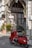 A vibrant red scooter is parked on a cobblestone street in front of a charming old building with an arched entrance. The facade is adorned with potted plants and a wooden lattice door. Signs with 'No Parking' and menu items are visible, adding a quaint touch to the setting.