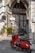 A vibrant red scooter is parked on a cobblestone street in front of a charming old building with an arched entrance. The facade is adorned with potted plants and a wooden lattice door. Signs with 'No Parking' and menu items are visible, adding a quaint touch to the setting.