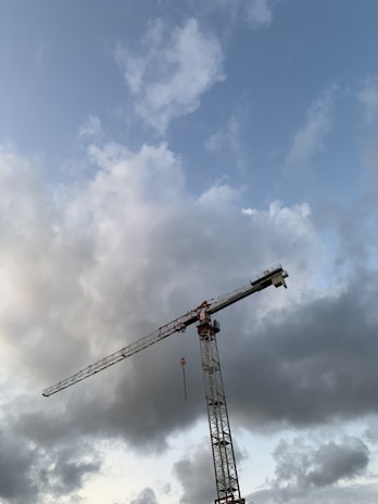 A towering construction crane extends into a mostly cloudy sky, with some patches of blue visible. The clouds appear thick and white with some darker areas, suggesting an overcast weather condition. The crane is positioned towards the right side of the image, highlighting its intricate metal framework.