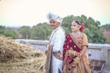A couple dressed in traditional Indian wedding attire is standing outdoors. The man is wearing a white sherwani with intricate designs and a white turban adorned with gold embellishments. The woman is wearing a richly decorated maroon bridal lehenga with heavy jewelry including a nose ring, necklace, and bangles. They are standing near a stack of hay with greenery and trees in the background.