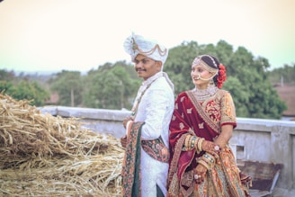 A couple dressed in traditional Indian wedding attire is standing outdoors. The man is wearing a white sherwani with intricate designs and a white turban adorned with gold embellishments. The woman is wearing a richly decorated maroon bridal lehenga with heavy jewelry including a nose ring, necklace, and bangles. They are standing near a stack of hay with greenery and trees in the background.