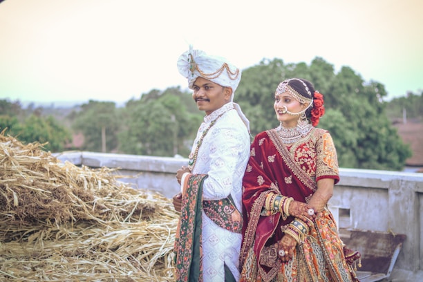 A couple dressed in traditional Indian wedding attire is standing outdoors. The man is wearing a white sherwani with intricate designs and a white turban adorned with gold embellishments. The woman is wearing a richly decorated maroon bridal lehenga with heavy jewelry including a nose ring, necklace, and bangles. They are standing near a stack of hay with greenery and trees in the background.