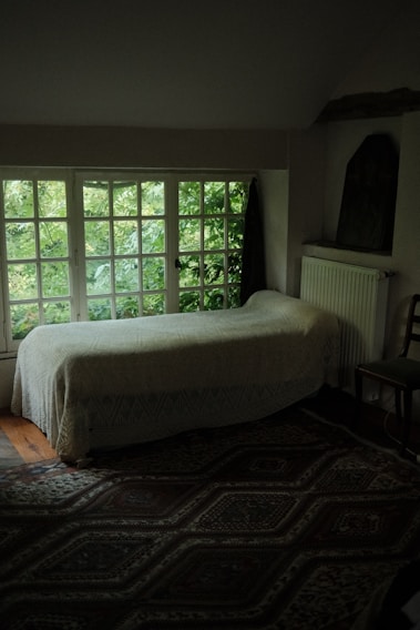 A cozy room with a single bed covered in a light quilt positioned against a wall with a large window showing lush green foliage outside. The room is dimly lit with shadows on the intricately patterned rug on the floor. A radiator is mounted on the wall next to a simple wooden chair.