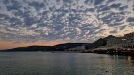 A serene waterfront scene at dusk with scattered clouds illuminated by the fading sunlight. The shoreline is lined with buildings, including a hotel with a sign, while the calm water reflects the evening sky. Hills silhouette in the background add depth to the horizon.