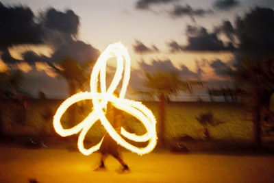 A person is performing a fire dance at dusk, creating intricate patterns with bright flames. The background features a beach scene with palm trees and a colorful sky filled with dark clouds as the sun sets.