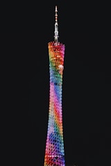 A tall, slender tower illuminated by a vibrant display of rainbow-colored lights against a dark night sky. The structure features a lattice design with a tapering shape, culminating in an antenna at the top.