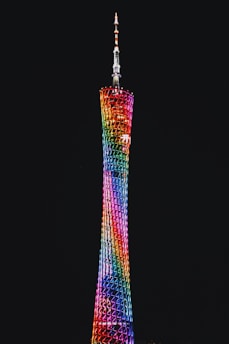 A tall, slender tower illuminated by a vibrant display of rainbow-colored lights against a dark night sky. The structure features a lattice design with a tapering shape, culminating in an antenna at the top.