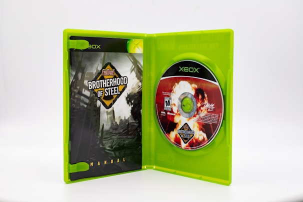 An open green Xbox game case containing a manual on the left side, titled 'Fallout Brotherhood of Steel', with a dark post-apocalyptic design. On the right side, a game disc is inserted, featuring vivid imagery including an explosion and the same title.