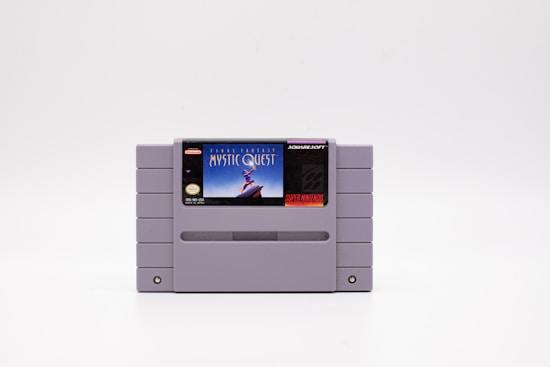 A Super Nintendo Entertainment System (SNES) game cartridge featuring the game 'Final Fantasy Mystic Quest.' The cartridge is primarily gray with a label on the front displaying the game's title and artwork. The label features an illustration of a heroic character standing on a cliff against a blue sky.