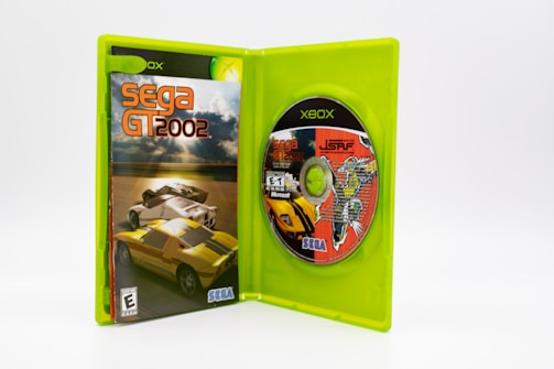 An open green Xbox game case displays the cover and disc of the Sega GT 2002 game. The cover features two sports cars racing on a track under a cloudy sky. The disc includes colorful graphics and text, indicating compatibility details and featuring another game, Jet Set Radio Future.