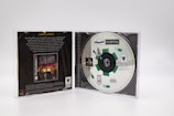 A PlayStation game case is open, revealing the disc for 'Theme Hospital' on the right side. The disc features a design with green pills and a mechanical theme. The left side shows the back cover of a different game, 'Warhammer: Dark Omen,' with promotional text and graphics.