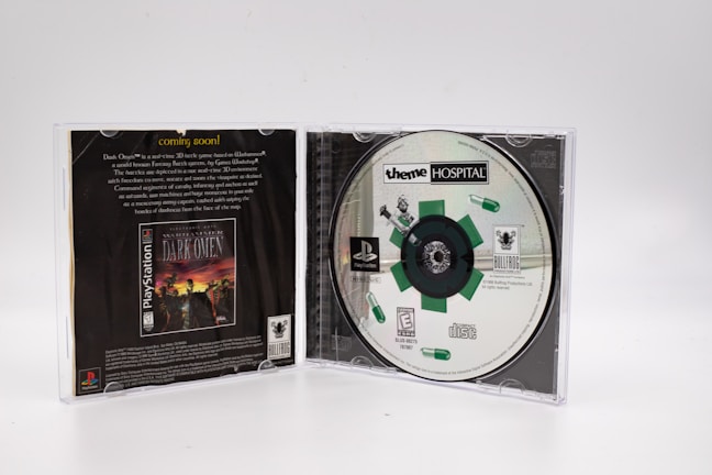 A PlayStation game case is open, revealing the disc for 'Theme Hospital' on the right side. The disc features a design with green pills and a mechanical theme. The left side shows the back cover of a different game, 'Warhammer: Dark Omen,' with promotional text and graphics.