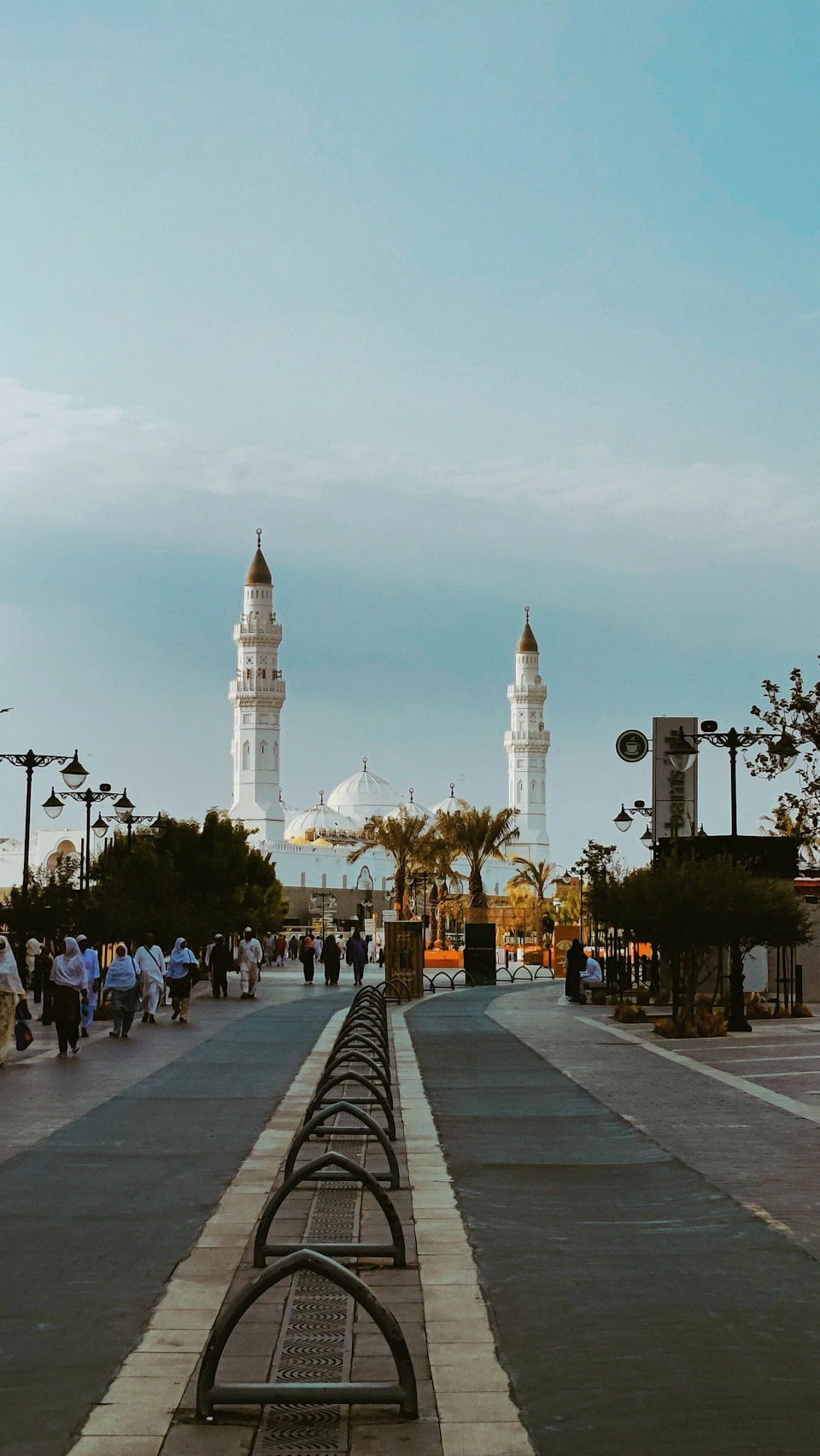The image depicts a bustling city scene with a large mosque featuring two towering minarets and a dome, surrounded by lush greenery. The mosque is situated on a street lined with trees and lampposts, creating an urban oasis amidst the natural surroundings. A sidewalk runs through the center of the frame, where several people can be seen walking or standing, adding to the lively atmosphere.