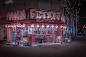 A cozy Japanese izakaya with traditional lanterns illuminating the exterior. Two people are seated at an outdoor table, enjoying their meals. The establishment is adorned with wooden panels, red accents, and vertical banners featuring Japanese characters. The street is quiet and dimly lit, with electrical wires visible against the night sky.