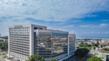 A modern, large office building with a glass facade standing prominently in an urban area. The architecture is sleek and geometric, and the surroundings include several smaller buildings and tree-lined streets. The sky is clear and blue, indicating a bright day.