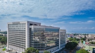 A modern, large office building with a glass facade standing prominently in an urban area. The architecture is sleek and geometric, and the surroundings include several smaller buildings and tree-lined streets. The sky is clear and blue, indicating a bright day.
