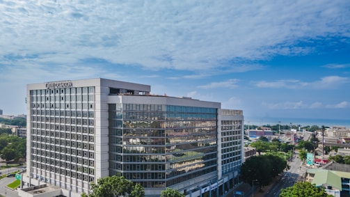 A modern, large office building with a glass facade standing prominently in an urban area. The architecture is sleek and geometric, and the surroundings include several smaller buildings and tree-lined streets. The sky is clear and blue, indicating a bright day.