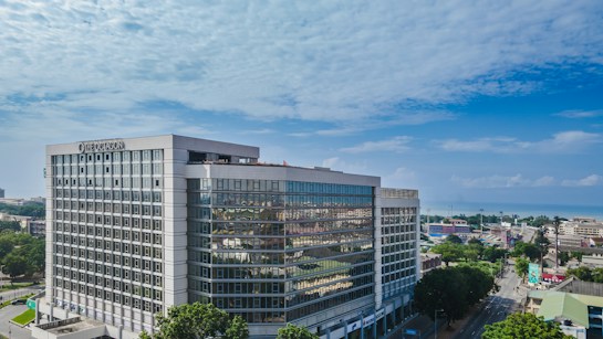 A modern, large office building with a glass facade standing prominently in an urban area. The architecture is sleek and geometric, and the surroundings include several smaller buildings and tree-lined streets. The sky is clear and blue, indicating a bright day.