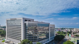 A modern, large office building with a glass facade standing prominently in an urban area. The architecture is sleek and geometric, and the surroundings include several smaller buildings and tree-lined streets. The sky is clear and blue, indicating a bright day.