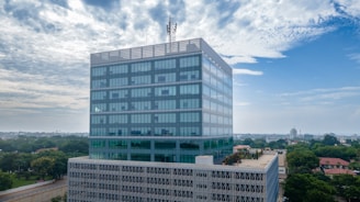 A modern, multi-story office building with reflective glass windows stands prominently against a partly cloudy sky. The structure appears to have a rooftop garden or terrace with some visible greenery. The surrounding area is composed of trees and other buildings, suggesting an urban environment.