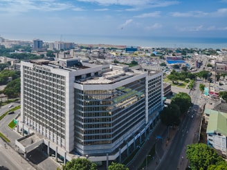 An aerial view of a large modern office building situated in an urban area. The building has a rectangular form with multiple floors and is surrounded by tree-lined streets and other smaller structures. In the background, there is an expansive view of a coastline with the sea meeting the horizon. The sky is mostly clear with a few scattered clouds.