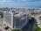 An aerial view of a large modern office building situated in an urban area. The building has a rectangular form with multiple floors and is surrounded by tree-lined streets and other smaller structures. In the background, there is an expansive view of a coastline with the sea meeting the horizon. The sky is mostly clear with a few scattered clouds.