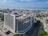 An aerial view of a large modern office building situated in an urban area. The building has a rectangular form with multiple floors and is surrounded by tree-lined streets and other smaller structures. In the background, there is an expansive view of a coastline with the sea meeting the horizon. The sky is mostly clear with a few scattered clouds.