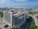 An aerial view of a large modern office building situated in an urban area. The building has a rectangular form with multiple floors and is surrounded by tree-lined streets and other smaller structures. In the background, there is an expansive view of a coastline with the sea meeting the horizon. The sky is mostly clear with a few scattered clouds.
