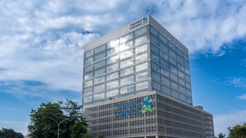 A modern office building with a reflective glass facade rises against a partly cloudy sky. The structure features a grid of windows and displays the logo and name of a financial institution near the top. Surrounding the building, there are some trees and streetlights, adding a touch of greenery to the urban setting.