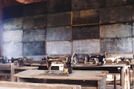 A classroom setting features several old-fashioned sewing machines on wooden desks lined in rows. The background includes a wall of chalkboards covered with writing, creating an atmosphere of a vintage educational or vocational training environment.