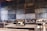 A classroom setting features several old-fashioned sewing machines on wooden desks lined in rows. The background includes a wall of chalkboards covered with writing, creating an atmosphere of a vintage educational or vocational training environment.