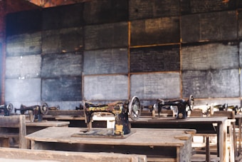 A classroom setting features several old-fashioned sewing machines on wooden desks lined in rows. The background includes a wall of chalkboards covered with writing, creating an atmosphere of a vintage educational or vocational training environment.