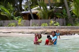 Children are playing in shallow ocean water near a sandy beach. They are in front of a fence with a tropical garden and palm trees in the background. Some children are standing and others appear to be swimming or wading in the water.