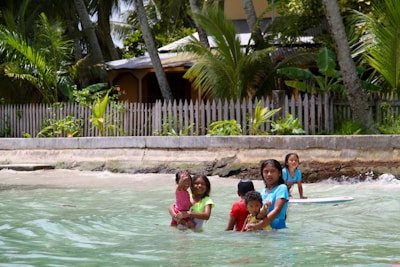 Children are playing in shallow ocean water near a sandy beach. They are in front of a fence with a tropical garden and palm trees in the background. Some children are standing and others appear to be swimming or wading in the water.