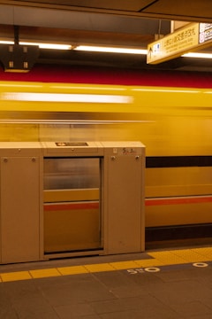 A subway station platform features a safety barrier in the foreground, designed with a small door, and markings on the floor. A train in motion passes by, creating a blurred effect, while overhead signs provide directions in multiple languages.