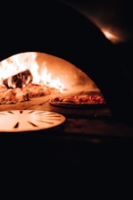 A wood-fired oven interior with a visible flame and a pizza being baked on the stone surface. The warm glow of the fire creates a cozy ambiance, and a round metal pizza peel is partially visible in the foreground.