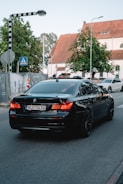 A black BMW sedan with the license plate NJ-1434 is driving down a street near an intersection. There is a pedestrian crossing sign to the left and a second car is visible in the background. The surroundings include a large tree and a building with a pitched roof and white facade.