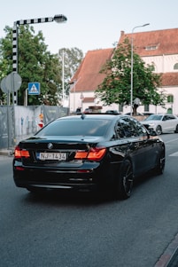 A black BMW sedan with the license plate NJ-1434 is driving down a street near an intersection. There is a pedestrian crossing sign to the left and a second car is visible in the background. The surroundings include a large tree and a building with a pitched roof and white facade.