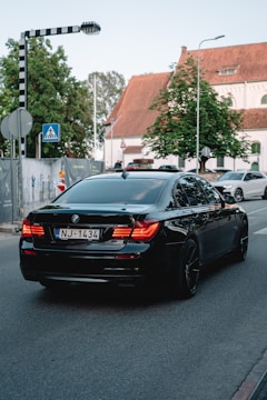A black BMW sedan with the license plate NJ-1434 is driving down a street near an intersection. There is a pedestrian crossing sign to the left and a second car is visible in the background. The surroundings include a large tree and a building with a pitched roof and white facade.