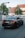 A black BMW sedan with the license plate NJ-1434 is driving down a street near an intersection. There is a pedestrian crossing sign to the left and a second car is visible in the background. The surroundings include a large tree and a building with a pitched roof and white facade.