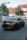 A black BMW sedan with the license plate NJ-1434 is driving down a street near an intersection. There is a pedestrian crossing sign to the left and a second car is visible in the background. The surroundings include a large tree and a building with a pitched roof and white facade.