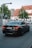 A black BMW sedan with the license plate NJ-1434 is driving down a street near an intersection. There is a pedestrian crossing sign to the left and a second car is visible in the background. The surroundings include a large tree and a building with a pitched roof and white facade.