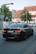 A black BMW sedan with the license plate NJ-1434 is driving down a street near an intersection. There is a pedestrian crossing sign to the left and a second car is visible in the background. The surroundings include a large tree and a building with a pitched roof and white facade.