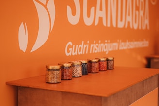 A group of small jars filled with different types of grains is neatly arranged on a wooden surface. The background features an orange wall with part of a large white logo and some text.