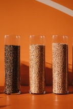 Four tall glass cylinders are filled with different types of grains and seeds, placed against a solid orange background. The contents range in color from dark brown to light beige, creating a contrast with the vibrant hue behind them.
