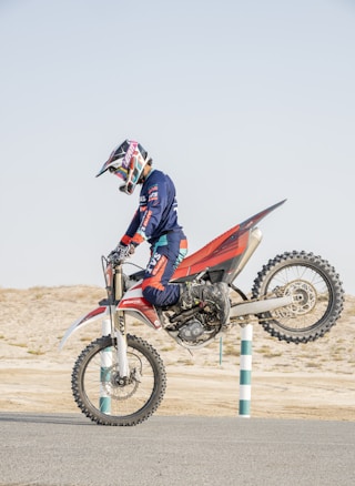 A motorcyclist is performing a wheelie on a dirt bike in a barren landscape. The rider is fully geared with a helmet, gloves, and protective clothing, showing a dynamic action pose with the front wheel lifted off the ground.