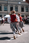 Several people are seen dancing in traditional attire on a street. Their costumes include red vests, white shirts, striped skirts, and decorated hairpieces. The background features a building with multiple windows and a clock above. There is a small crowd observing the performance.