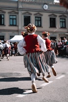 Several people are seen dancing in traditional attire on a street. Their costumes include red vests, white shirts, striped skirts, and decorated hairpieces. The background features a building with multiple windows and a clock above. There is a small crowd observing the performance.