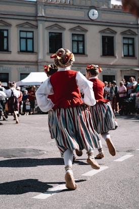 Several people are seen dancing in traditional attire on a street. Their costumes include red vests, white shirts, striped skirts, and decorated hairpieces. The background features a building with multiple windows and a clock above. There is a small crowd observing the performance.